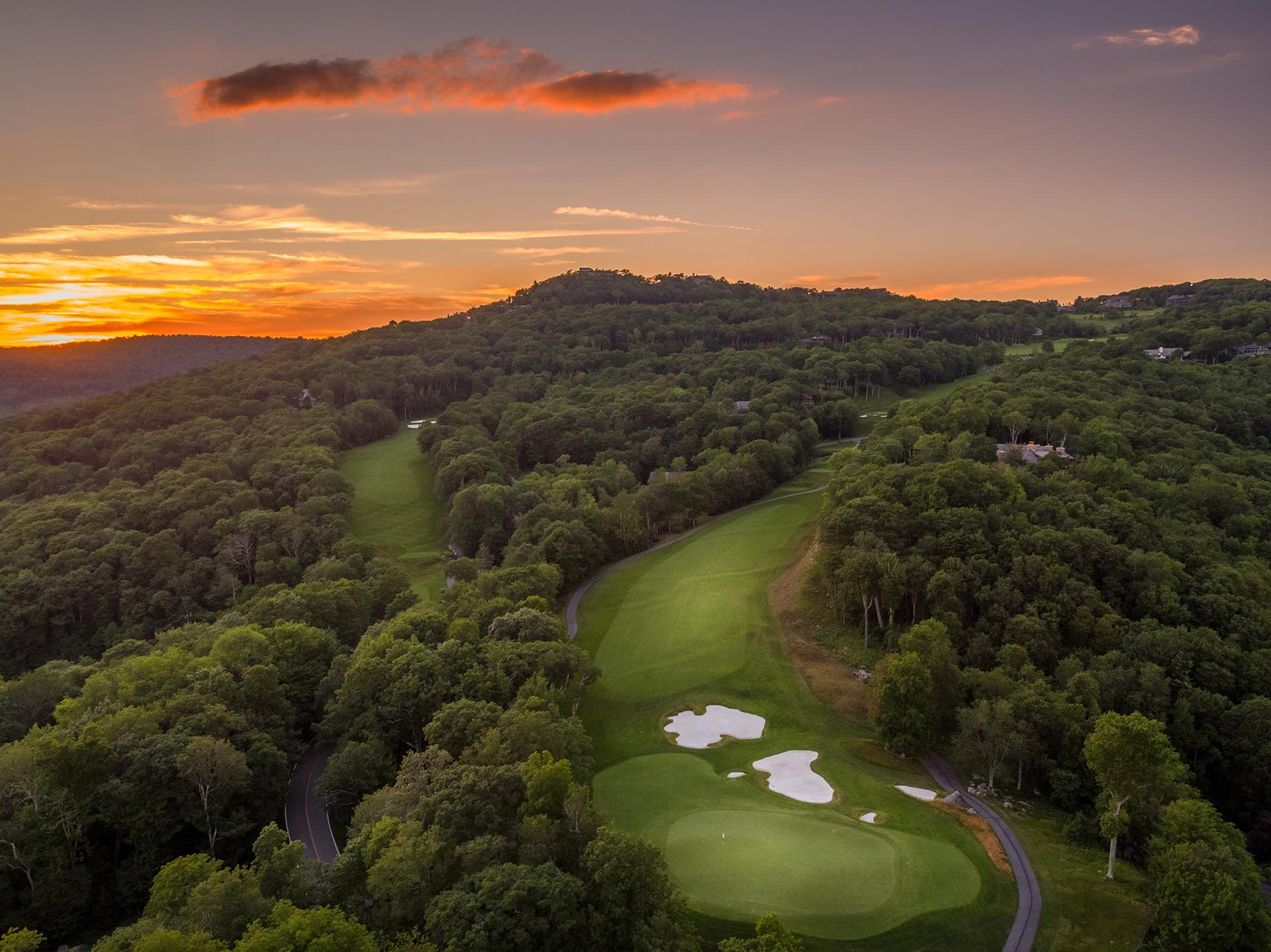 Aerial view of a forested golf course at sunset with vibrant orange sky.